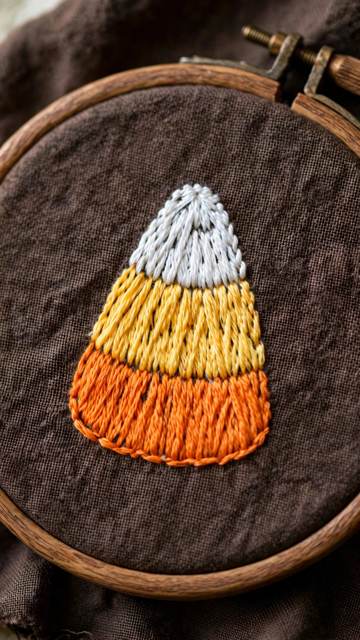 Photorealistic close-up of a candy corn hand-embroidered in orange, yellow and white thread on dark brown linen