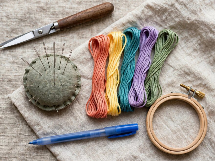 Beginner embroidery tools flat lay on linen: scissors, needles, floss skeins and a wooden hoop