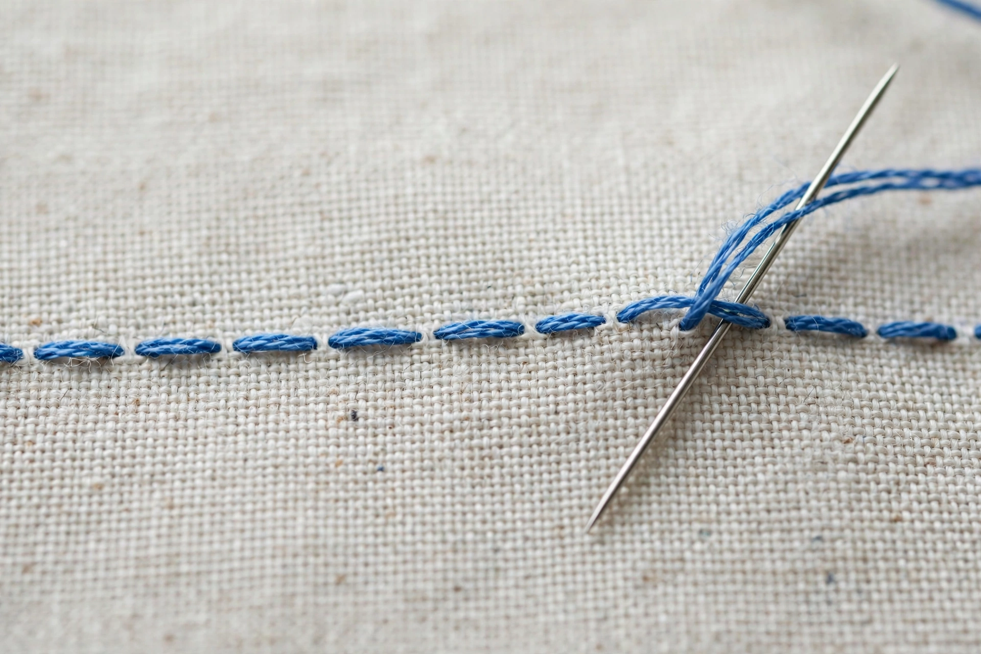 Macro close-up of a running stitch row in blue floss on white linen, needle mid-stitch, crisp thread detail, soft daylight