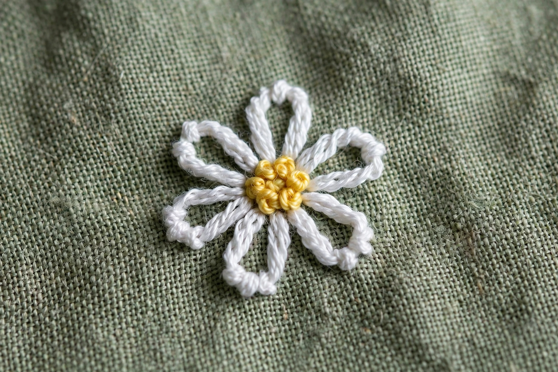 Close-up of lazy daisy stitches forming a white flower with a yellow french knot center on sage linen