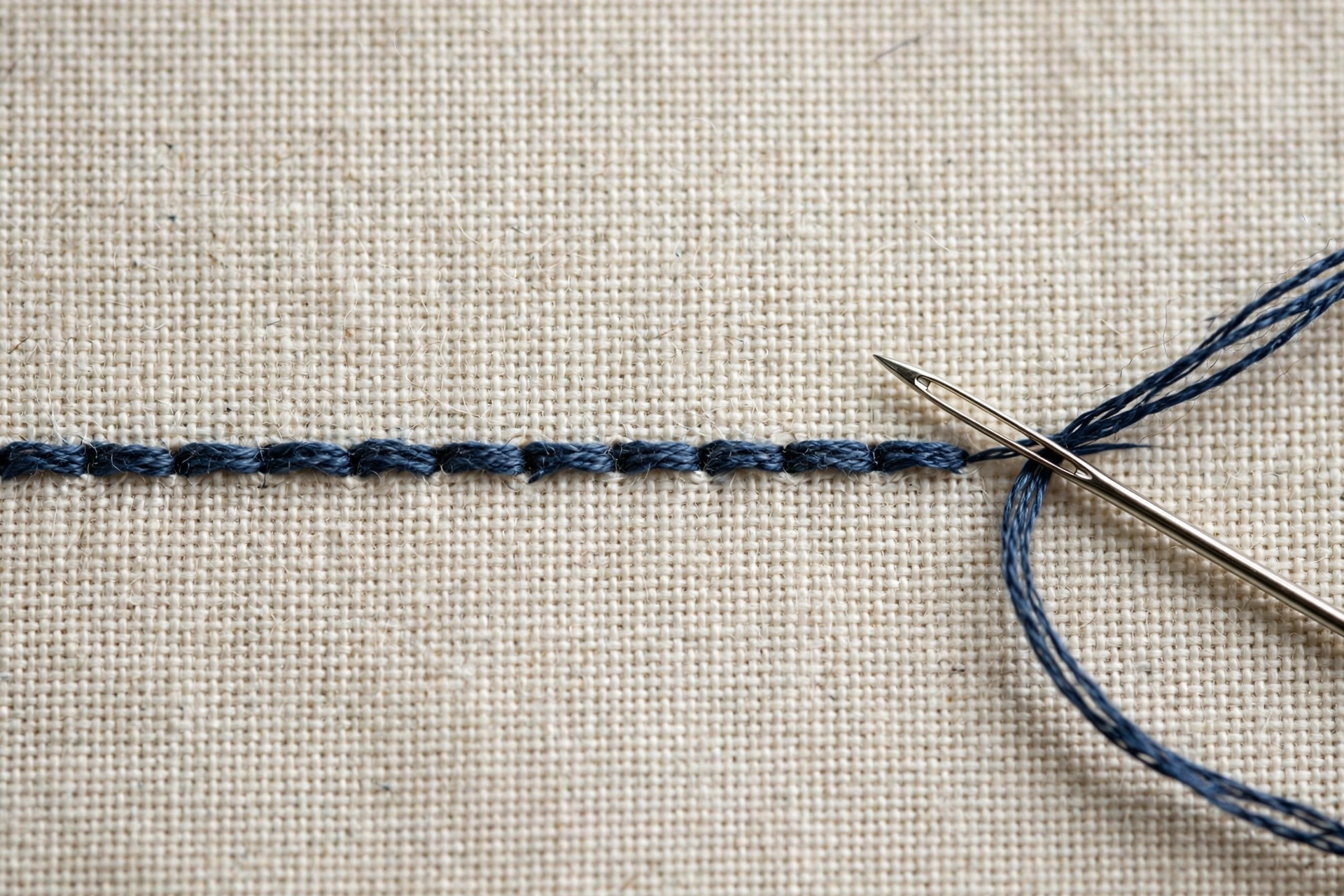 Macro close-up of backstitch worked in dark blue floss on cream linen, showing the even segmented line, needle in frame