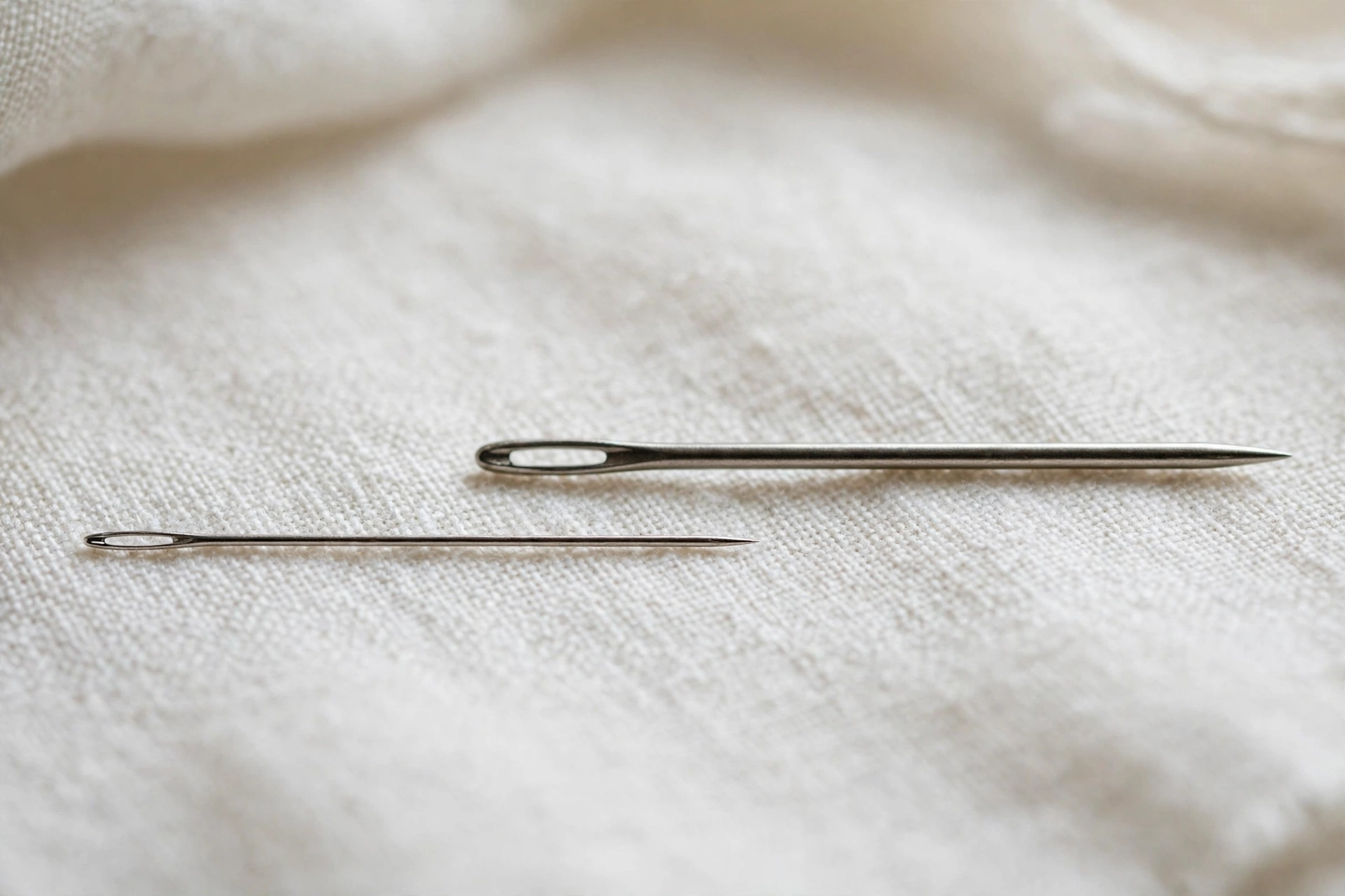 Macro shot of a sharp embroidery needle beside a crewel needle on white linen showing the slim shaft difference, shallow dept