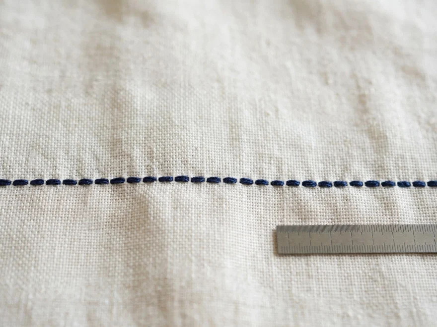 Macro close-up of perfectly even backstitch row in navy floss on white linen, small ruler beside showing consistent spacing