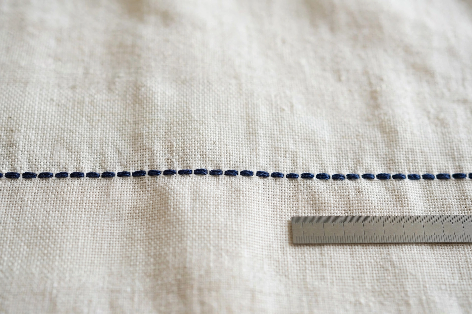 Macro close-up of perfectly even backstitch row in navy floss on white linen, small ruler beside showing consistent spacing