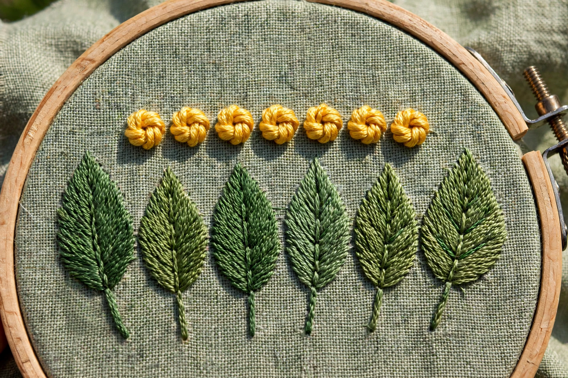 Row of even golden-yellow bullion knots stitched as flower stamens on green leaves, embroidery hoop edge visible, bright dayl
