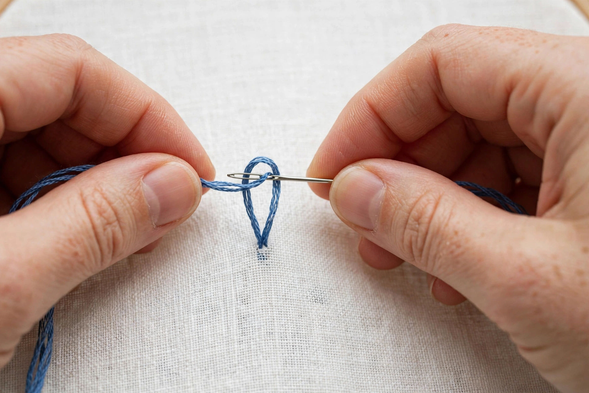 Photorealistic close-up of hands demonstrating the loop start, a loop of blue embroidery floss threaded through a silver needle over white linen, landscape orientation, no text