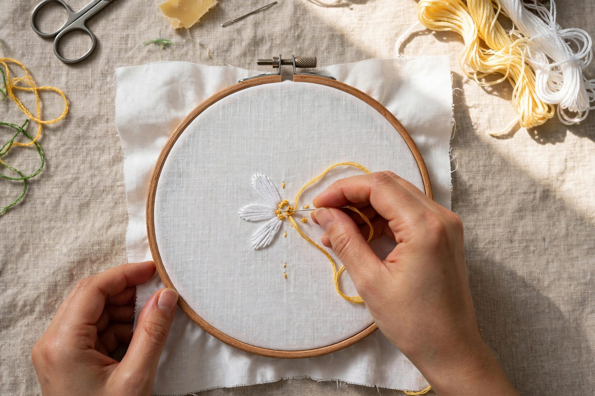 Close-up of hands stitching a pink rose on cream linen in a wooden hoop, soft daylight