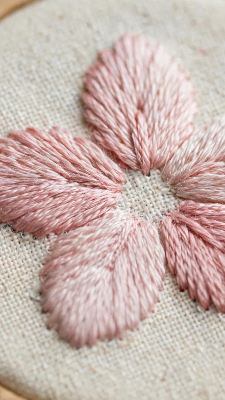 Close-up of satin stitch petals on cream fabric showing fine thread detail in a small hoop, no text