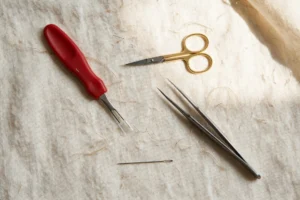 Photorealistic flat lay of embroidery frogging tools: seam ripper, small sharp embroidery scissors, tweezers, needle, on cream linen, bright daylight, landscape orientation, no text